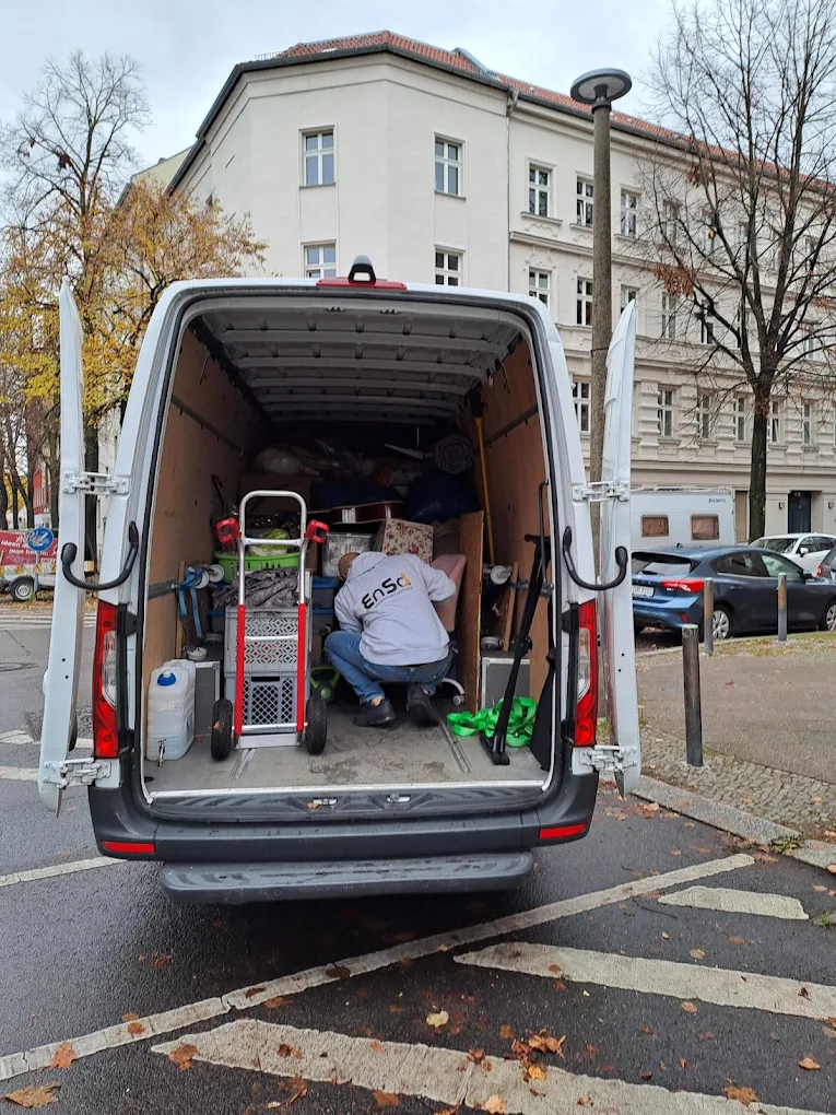 Person kniet im hinteren Teil eines offenen weißen Lieferwagens und ordnet Gegenstände wie eine Sackkarre und Kartons. Der Lieferwagen steht auf einer Stadtstraße, im Hintergrund sind Herbstbäume und Gebäude zu sehen.
