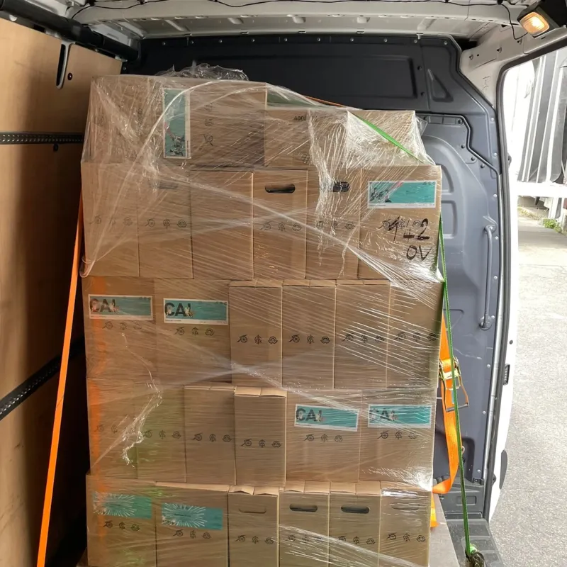 Stapel von mit Plastikfolie umwickelten Kartons, die mit orangefarbenen und grünen Gurten gesichert sind, im hinteren Teil eines Lieferwagens.