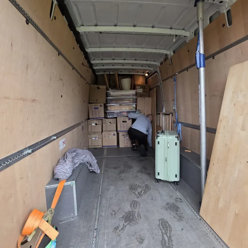 Innenraum eines Umzugswagens mit einer Person, die neben gestapelten Kisten und einem grünen Koffer auf der rechten Seite kauert.