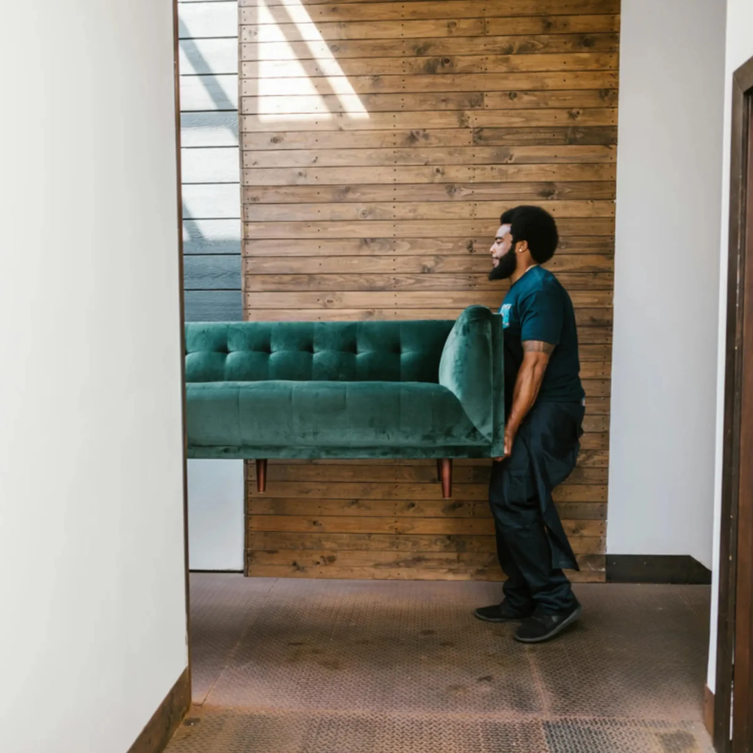 Mann mit Bart und Afro, der eine grüne Samtcouch vor einer Holzwand ins Haus trägt.